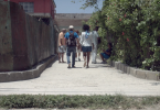 Zaima y Tomás caminan juntos por un pasillo estrecho en su barrio, rodeados de otras personas, vegetación y estructuras deterioradas.